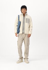 Creme fleece jakke med grå detaljer, fuld lynlås, sidelommer, sammen med beige cargo bukser og beige sneakers. Blå skuldertaske.