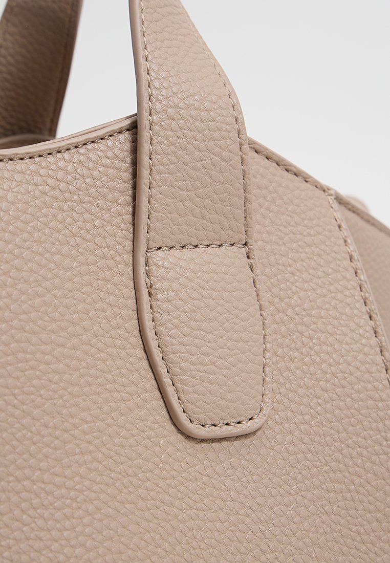 Primer plano de un bolso de cuero beige texturizado con detalles de costura en la correa, sobre un fondo liso.