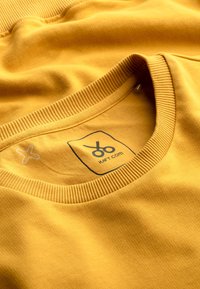 Felpa girocollo in cotone giallo senape, con colletto e orlo a costine. Presenta un logo ricamato e un'etichetta di cura interna. Tessuto morbido.