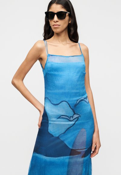 Μπλε, διαφανές φόρεμα με σχέδιο κύματος και πουλιού, λεπτές ρυθμιζόμενες τιράντες και αέρινη σιλουέτα. Συνδυάζεται με μαύρα γυαλιά ηλίου.