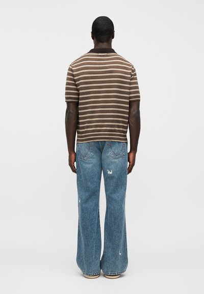 Άνδρας που στέκεται με την πλάτη γυρισμένη, φοράει ένα καφέ ριγέ κοντομάνικο πουκάμισο και φαρδιά μπλε τζιν με σκισίματα, μπροστά σε απλό φόντο.