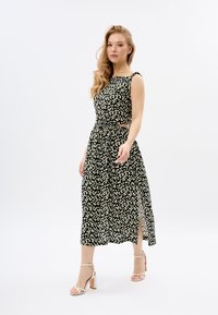 Schwarzes Blumenkleid mit weißen Gänseblümchen, ärmellos, tailliert, mit seitlichem Schlitz, knöchellang, kombiniert mit beigen Blockabsatzsandalen.