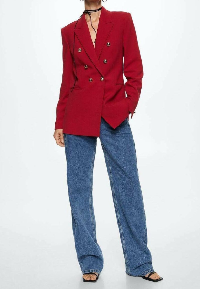 Femme portant un blazer rouge à double boutonnage, un jean bleu à coupe large, des sandales noires à talons et à brides, et un collier ras-du-cou noir.