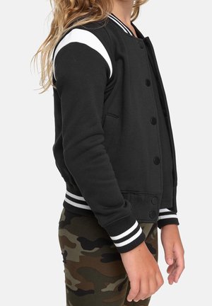 Zwart varsityjack met witte strepen op de schouders, drukknopen, geribbelde manchetten en zijzakken, gecombineerd met broek in camouflagepatroon.