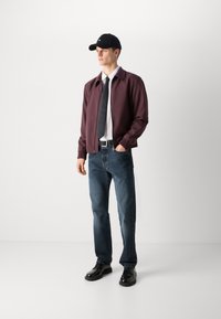 Bordo bomber-jakk, valge särk, must lips, tumedad teksad ja mustad kingad. Modell kannab musta mütsi ja seisab sirgelt.