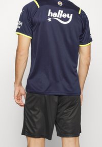 Laivaston sininen urheilupaita, jossa on keltaisia somisteita ja logo, lyhyet hihat, yhdistettynä mustiin shortseihin, joissa on hienovarainen tekstuuri ja kevyt muotoilu.