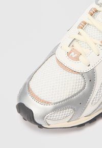 Sneaker bianca in rete con accenti argentati, dettagli beige e suola con texture. Presenta un design con lacci e logo sulla linguetta.