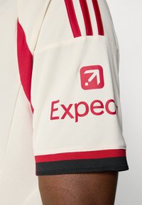 Kreemikollane jersööp punaste aktsentidega, millel on õlal kolm punast triipu ja silmapaistev punane "Expect" logo varrukal. Must alläär.