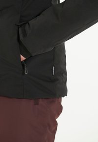 Svart vattentät jacka med en ficka med dragkedja, elastiska ärmslut och ett mjukt innerfoder, tillsammans med vinröda byxor.