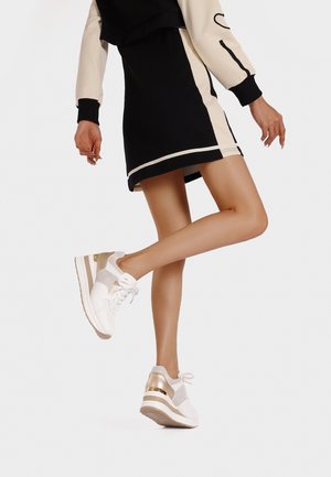 Sneakers bianchi con dettagli metallici dorati, rete testurizzata e una suola spessa. Ambientati su uno sfondo di un outfit nero e crema.