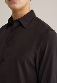 Črna srajca z gumbki in ovratnikom, ki se ponaša z gladko teksturo in diskretnim sijajem. Podrobnosti vključujejo gumbe spredaj in strukturirana ramena.