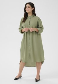 Abito camicia lungo e button-up in verde oliva, realizzato in tessuto morbido. Presenta un colletto, maniche arrotolabili e spacchi laterali.