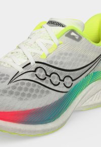 Λευκό αθλητικό παπούτσι Saucony με μαύρο λογότυπο, νέον κίτρινες λεπτομέρειες και τιρκουάζ-κόκκινο διαβαθμισμένο σχέδιο στην πλευρά της σόλας.