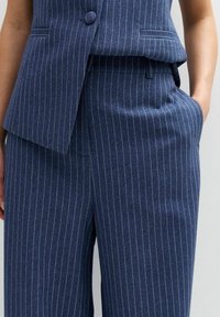 Costume bleu marine à rayures fines comprenant un gilet ajusté avec un bouton et un pantalon taille haute, fabriqué dans un tissu lisse avec des rayures verticales.