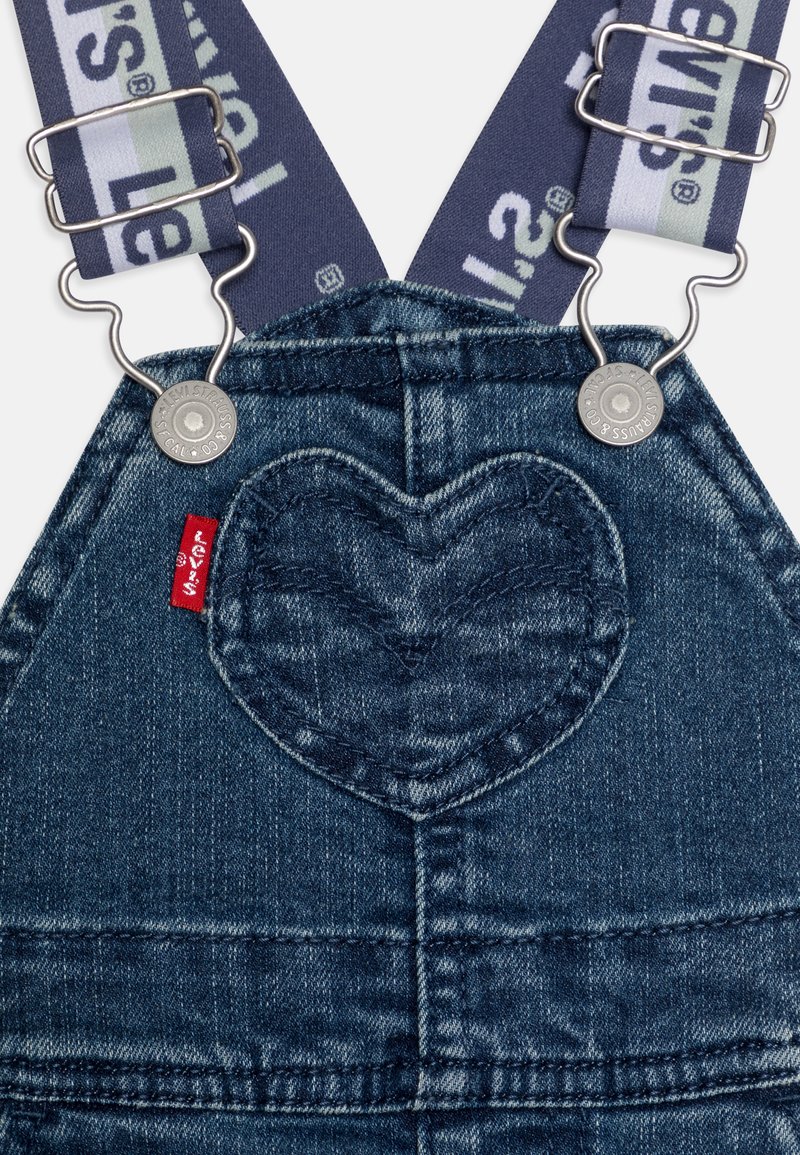 Denim-Latzhose mit herzförmigen Taschen, marineblauen Trägern mit einem Logo und silbernen Hardware-Details. Texturiert blauer Stoff.