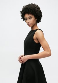 Μαύρο φόρεμα χωρίς μανίκια με εφαρμοστό μπούστο και φουσκωτή φούστα. Ομαλή υφή και μινιμαλιστικός σχεδιασμός, που αναδεικνύει καθαρές γραμμές και απλή σιλουέτα.