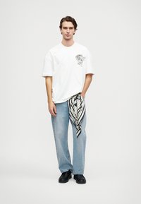 Λευκό κοντομάνικο μπλουζάκι με γραφικό σχέδιο, ανοιχτό μπλε τζιν και μία μαύρη-λευκή ριγέ γραβάτα δεμένη στη μέση. Μαύρα παπούτσια.