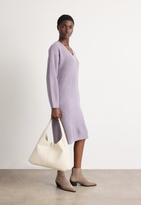 Gai violetā adīta kleita ar V-veida kakla izgriezumu un garām Piedurknēm, apvienota ar krēmkrāsas rokassomu un pelēkbrūnām potīšu zābaciņām ar blokveida papēdi.