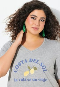 Szara koszulka typu V-neck z niebieskim napisem "COSTA DEL SOL" i grafiką cytryny. Modelka nosi zielone wiszące kolczyki i ma kręcone włosy.