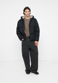 Jachetă puf din negru cu glugă gri, tricou bej și blugi negri largi. Prezintă un design simplu cu accente minime.