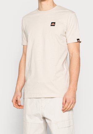 Mężczyzna w jasnobeżowej koszulce z krótkim rękawem z małym czarno-pomarańczowym logo na klatce piersiowej i rękawie, ubrany w jasne szorty bojówki.