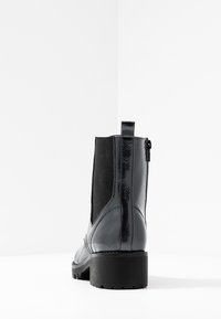 Botines de charol negro con paneles laterales elásticos, cremallera trasera y un tacón grueso y texturizado. Acabado suave y brillante con color sólido.