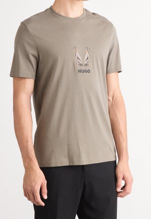 Mies, joka pukeutunut oliivinvihreään T-paitaan, jossa on kirjailtu kaksi koiraa ja teksti "HUGO", yhdistettynä mustiin housuihin, yksinkertaisen valkoisen taustan edessä.