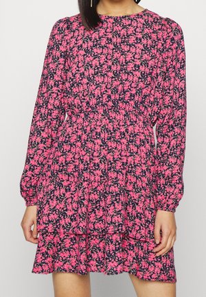 Femme portant une robe fleurie rose à manches longues avec une taille élastique et une jupe à volants, debout devant un fond clair uni.