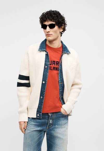 Jeune homme aux cheveux bouclés portant des lunettes de soleil noires, un cardigan crème tricoté avec des rayures noires, une veste en jean, un T-shirt rouge imprimé et un jean délavé.