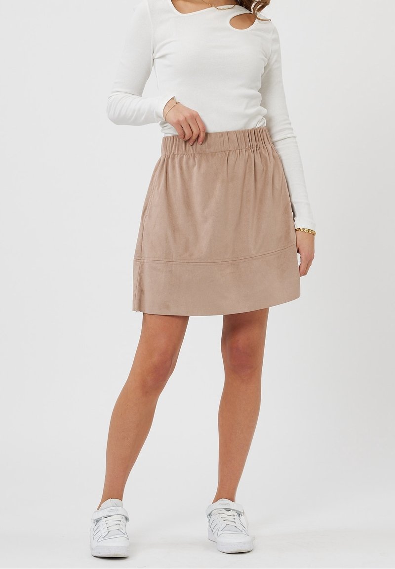 Femme portant une jupe beige trapèze à taille élastique, un haut blanc à manches longues avec découpe à l'épaule, des baskets blanches, un bracelet et un collier.