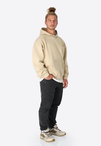 Beige Kapuzenpulli mit einer vorderen Tasche, kombiniert mit schwarzen Jeans und beigen Sportschuhen mit schwarzen Streifen. Das Modell steht frontal.