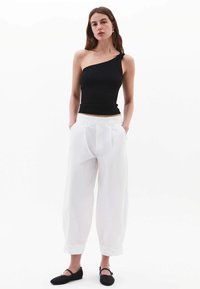 Top nero a spalla singola in materiale stretch aderente; abbinato a pantaloni bianchi a vestibilità ampia con pieghe e orli arrotolati; scarpe nere basse.