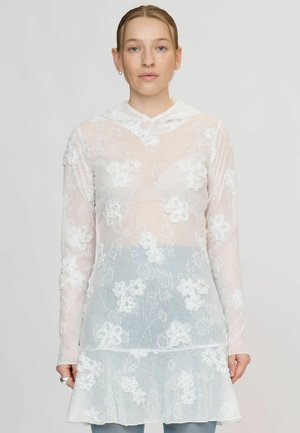 Jeune femme portant un haut à manches longues en dentelle florale blanche et transparente avec un col, debout devant un fond clair uni.