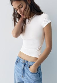 Fiatal nő hosszú sötét hajjal, fehér, testhez simuló pólót és világoskék farmert visel, egyik kezét a zsebében tartja, és lefelé néz.