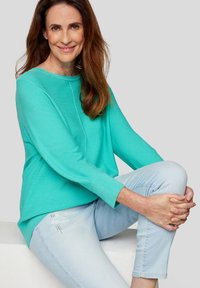 Türkiser Pullover mit langen Ärmeln und gerippten Bündchen, kombiniert mit hellblauen Jeans mit dezenten Used-Details, sitzende Pose.