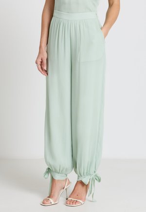 Pantalon vert clair à jambes larges avec des bas recueillis noués en nœuds aux chevilles, porté avec des sandales blanches à talons hauts et les mains dans les poches.