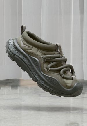 Αθλητικό παπούτσι σε ανοιχτό ελιά χρώμα με παχιά, υφή σόλα και κορδόνια σε βρόχο, αιωρούμενο πάνω από γυαλιστερό μαρμάρινο πάτωμα και λευκές κατακόρυφες περσίδες.