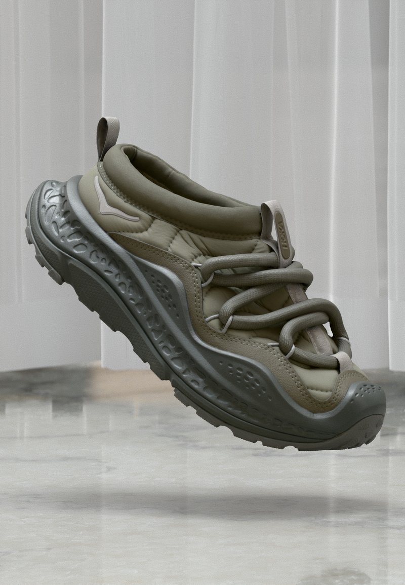 Αθλητικό παπούτσι σε ανοιχτό ελιά χρώμα με παχιά, υφή σόλα και κορδόνια σε βρόχο, αιωρούμενο πάνω από γυαλιστερό μαρμάρινο πάτωμα και λευκές κατακόρυφες περσίδες.