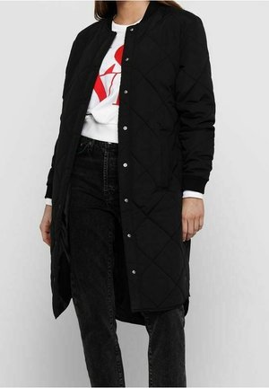 Manteau long matelassé noir avec fermeture à boutons-pression, col côtelé et manches longues. Porté sur un sweat-shirt blanc et un jean noir.