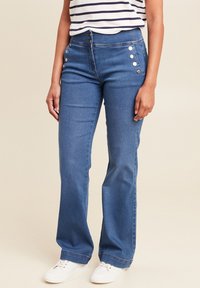 Blauwe flare jeans met een hoge taille, voorzien van metalen knoopaccenten aan de zijkant en een soepele denimtextuur. Gecombineerd met witte sneakers.