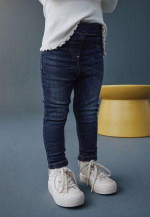 Kind trägt dunkelblaue Jeans, weißes Langarmshirt und weiße Turnschuhe und steht drinnen auf einem blauen Boden neben einem gelben Hocker.