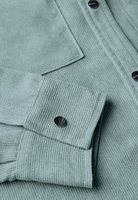 Světle modrá pruhovaná látková košile s černými patentkami a kapsou viditelnou poblíž manžety a přední légy.