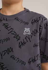 Siva majica s crnim, stiliziranim tekstom "SALTYDOG" i malim bijelim grafičkim prikazom buldoga na lijevoj strani prsnog područja. Kratki rukavi, dizajn s okruglim izrezom.