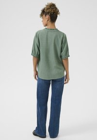 Blusa verde, con texture e maniche corte a volantino abbinata a jeans blu a gamba dritta. L'outfit include scarpe nere.