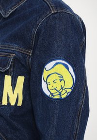 Denimjakke med gult brodert "M" og en rund lapp med en blå og gul tegneseriefigur på venstre arm.