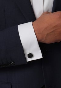 Navy suit arm with a fitted white cuff featuring a round black cufflink. Smooth texture with subtle pinstripes on the fabric.