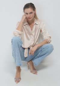 Donna con i capelli tirati indietro, che indossa una camicia di seta beige, jeans azzurri e scarpe col tacco beige, accovacciata contro un muro bianco.
