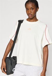 Svijetlo bež majica kratkih rukava s crvenim šavovima na ramenima i malim bež logotipom. Nosi se s sivim trenirkama i crnom torbom s bijelim detaljima.