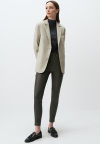 Beige Blazer über einem dunkelblauen gerippten Oberteil, kombiniert mit olivgrünen, figurbetonten Hosen und schwarzen Loafers mit dekorativen Quasten.