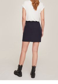 Woman with red hair wearing a white short-sleeve top, black mini skirt, and black lace-up combat boots, standing facing away on a plain background.